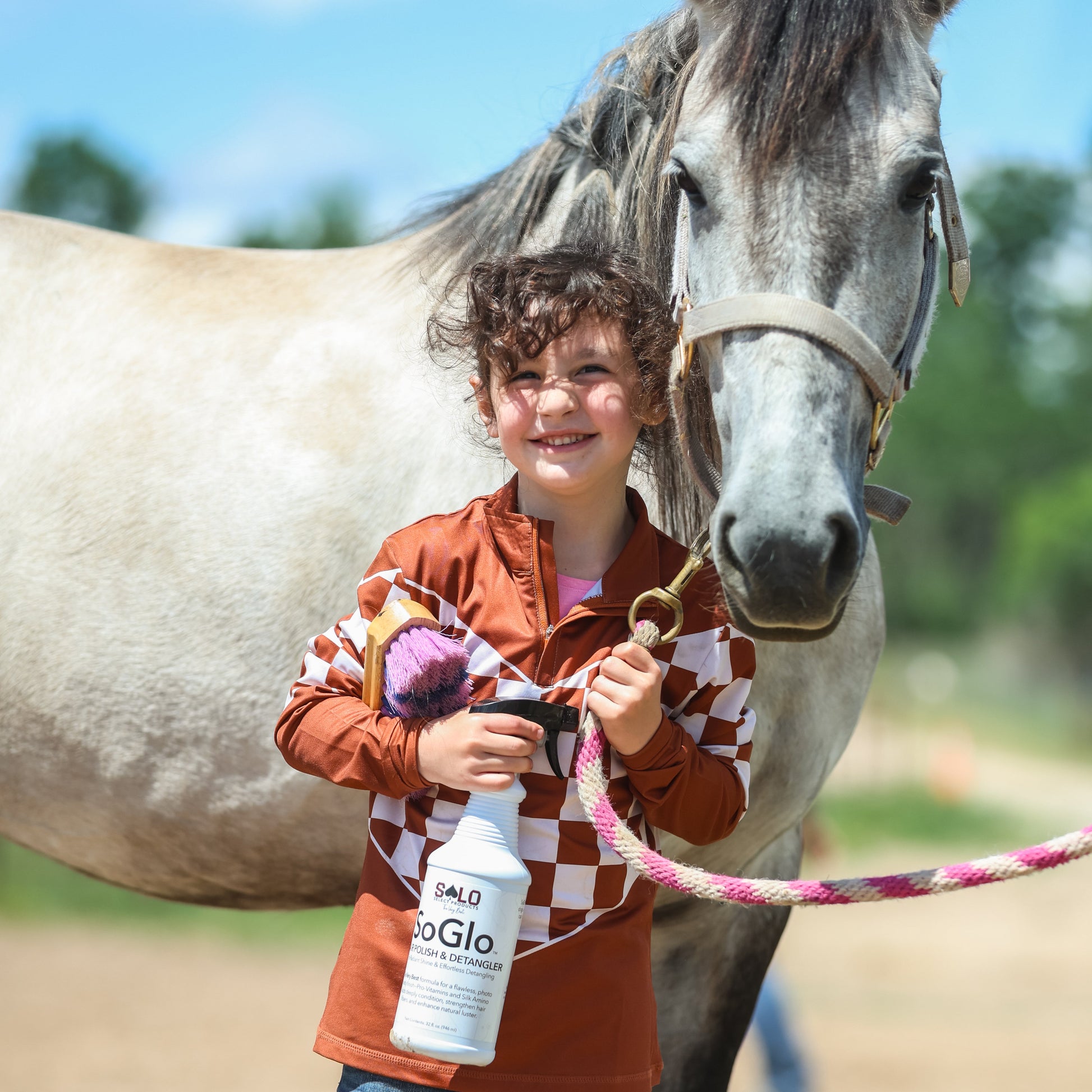 A child stands outdoors beside a horse, holding a white spray bottle labeled oGla. The child wears an orange and white checkered long-sleeve top with blue jeans, and the horse is in the background under a blue sky.